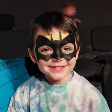 A little boy is smiling, his face is painted like batman.