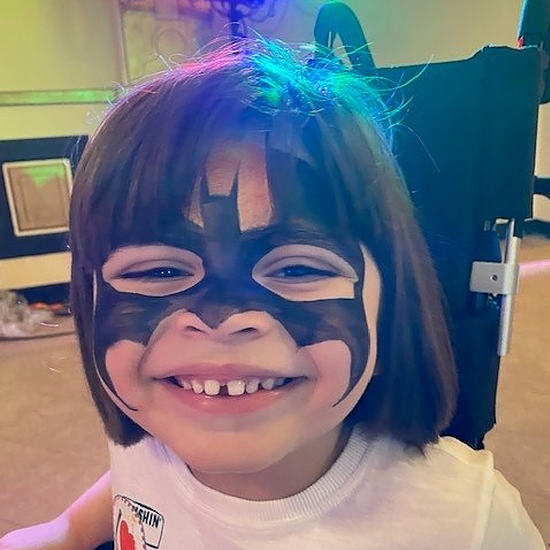 A little boy called Isaac is sat in his wheelchair at the circus show. He has bat man face paint, brown hair falling to just above his shoulders, and wears a circus starr sticker on his chest.