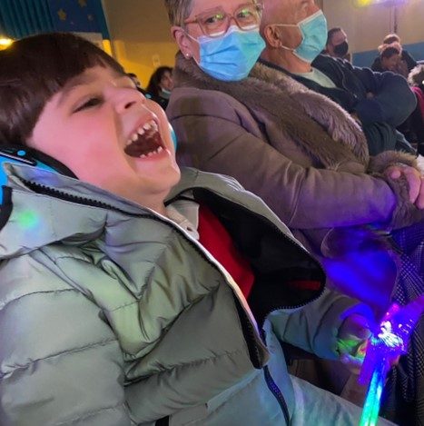 A little boy has his head back with laughter. He is wearing ear defenders and is holding a light up sensory wand. His Grandma is sat next to him looking at him whilst he laughs.