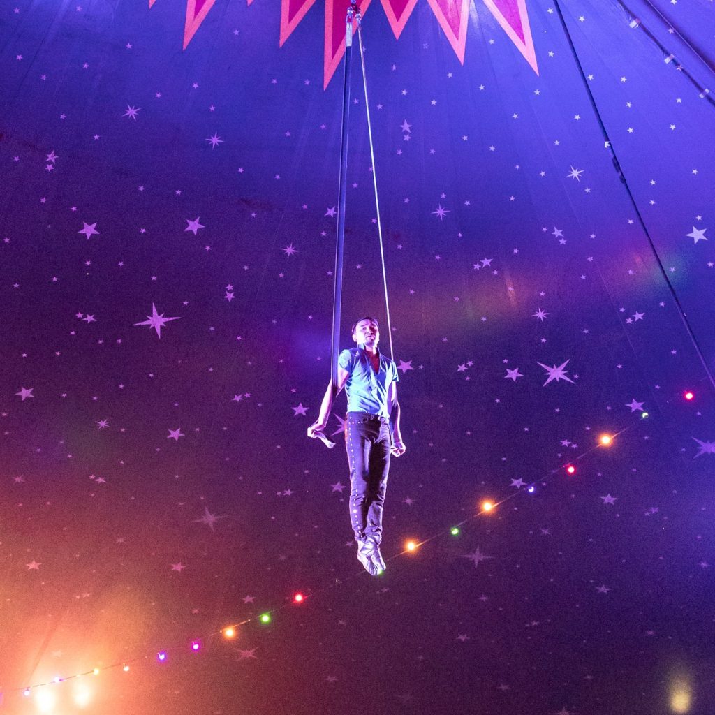 The image looks up towards the top of the big top, which has a blue canopy with white stars in different sizes. Hanging from the top of the tent are two blue straps. There is a man (named Ashkat) holding himself in the air on the straps. He has black trousers with diamantes running along the seam and a silky blue shirt. 