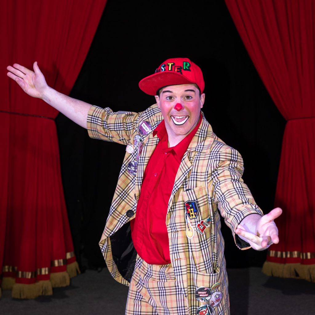 The clown is standing in front of the red circus curtains with his arms open and a big smile on his face. He has face make up that consists of a red nose, and white bottom lip. He is wearing a red cap that has his name 'Buster' written on it in different coloured bold letters. He is wearing tartan suit that has various different badges attached to his blazer.