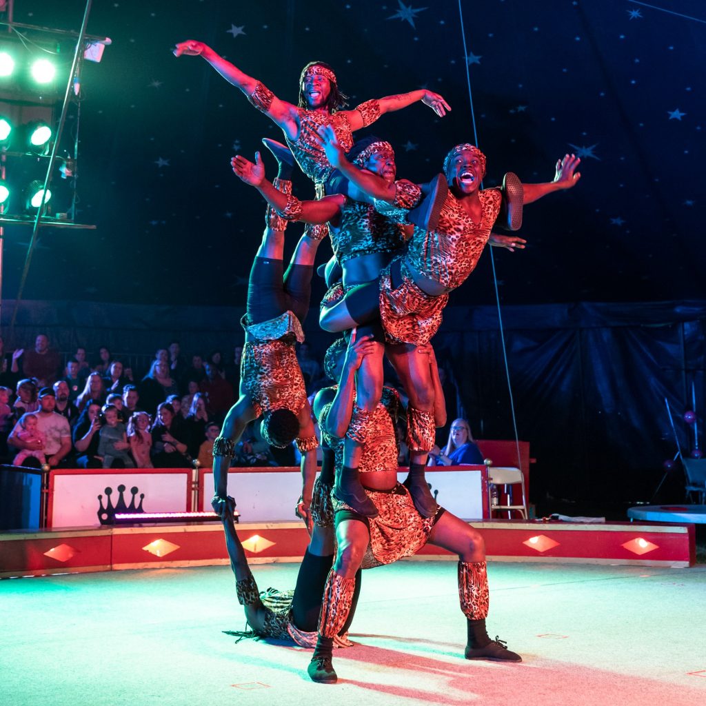 A group of acrobatics are balancing on each other inside of the circus ring. There are six men that are holding each other in different positions, it makes the man at the top of the group is waving his arms in the air like he has wings. The audience are sat in the seats around the ring.