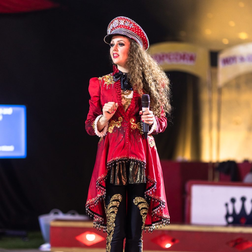 The ringmistress is stood in the circus ring. She is holding a microphone in her left hand. She is wearing a long red coat that featured gold appliques. She has long brown curly hair and a red hat that features silver diamantes. 