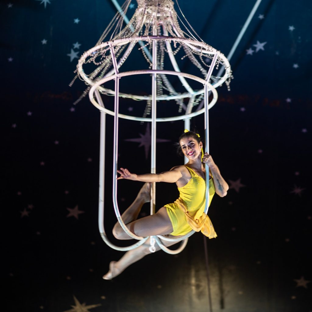 A woman (named Rita) is hanging on a large white chandelier high up in the roof of the big top tent. The chandelier has delicate strands of jewels hanging from the top. The woman has brown hair featuring a yellow bow and is wearing a yellow leotard with material handing from it. 