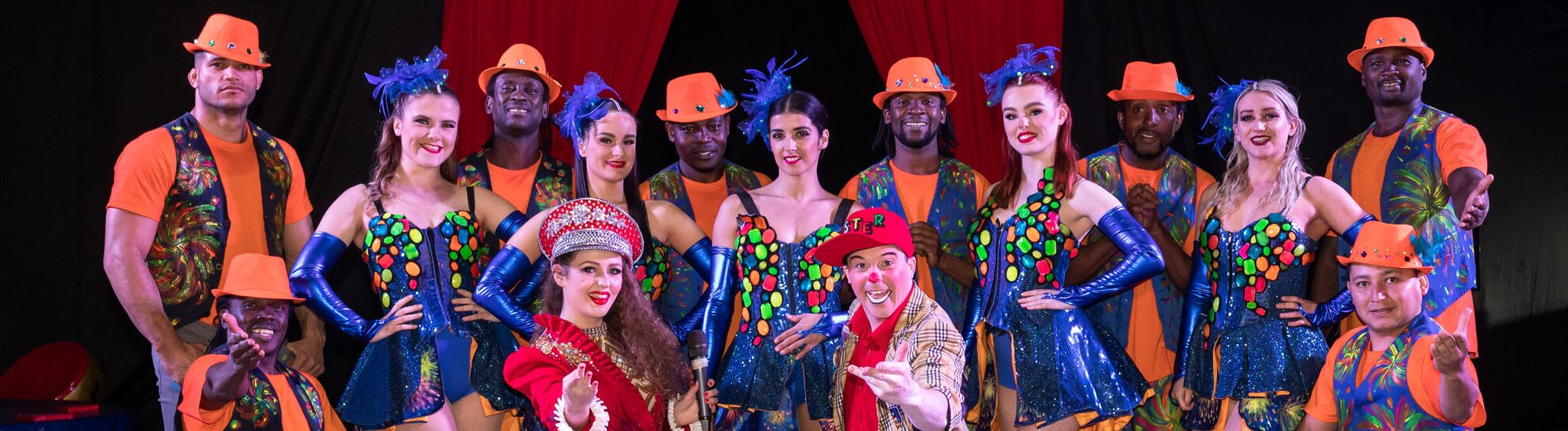A circus troupe pose in front of the circus ring curtains. There are both men and women wearing bright and colourful clothing.