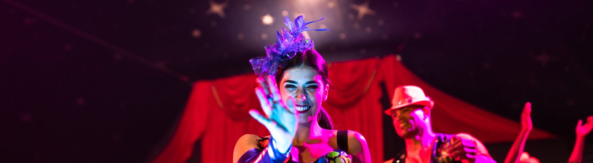 A dancer with brown hair waves at the camera. You can see the circus curtains in the background.