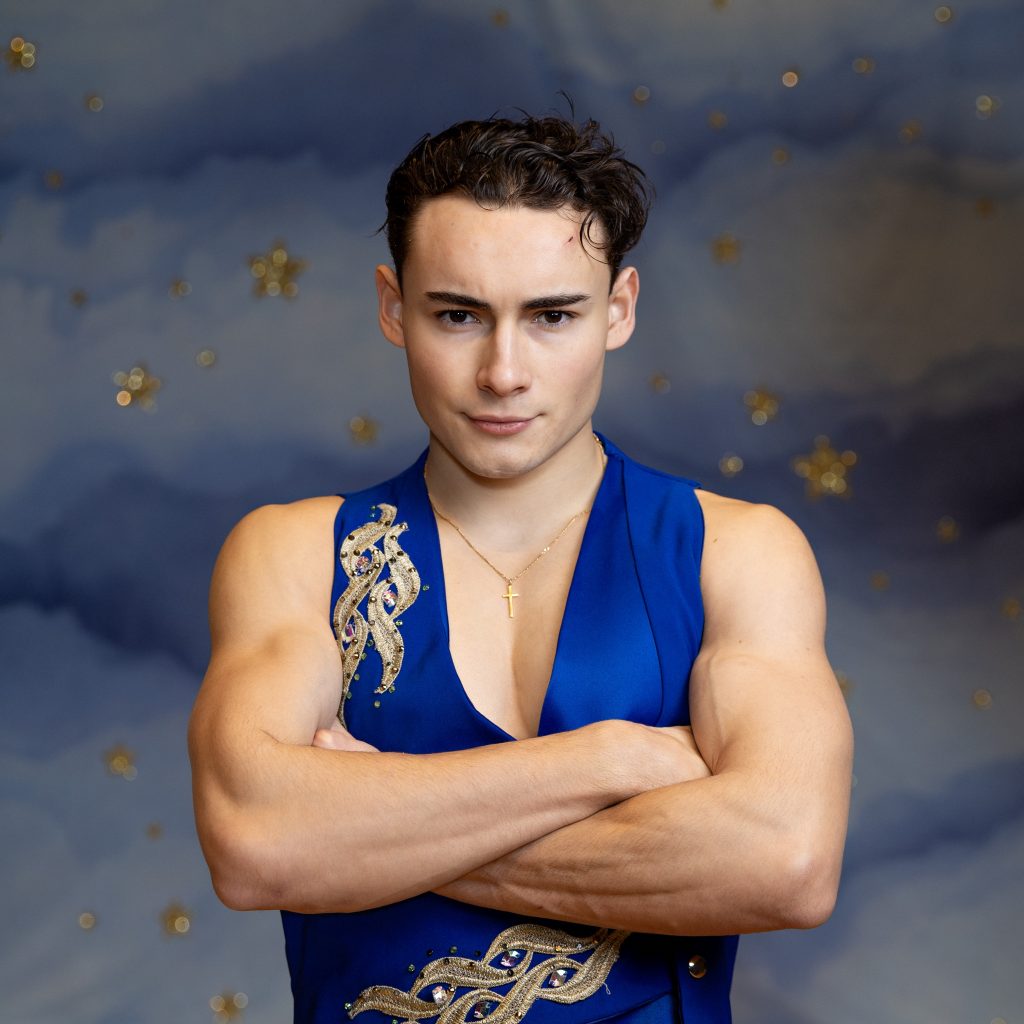 A man has his arms crossed, he is wearing a blue waistcoat with gold applique. 