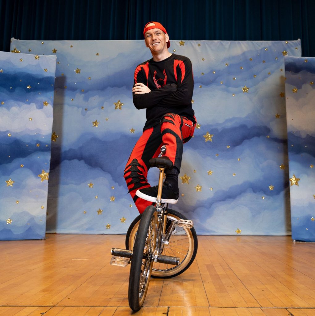 A man is sitting on a BMX bike, he is wearing a red and black outift. 