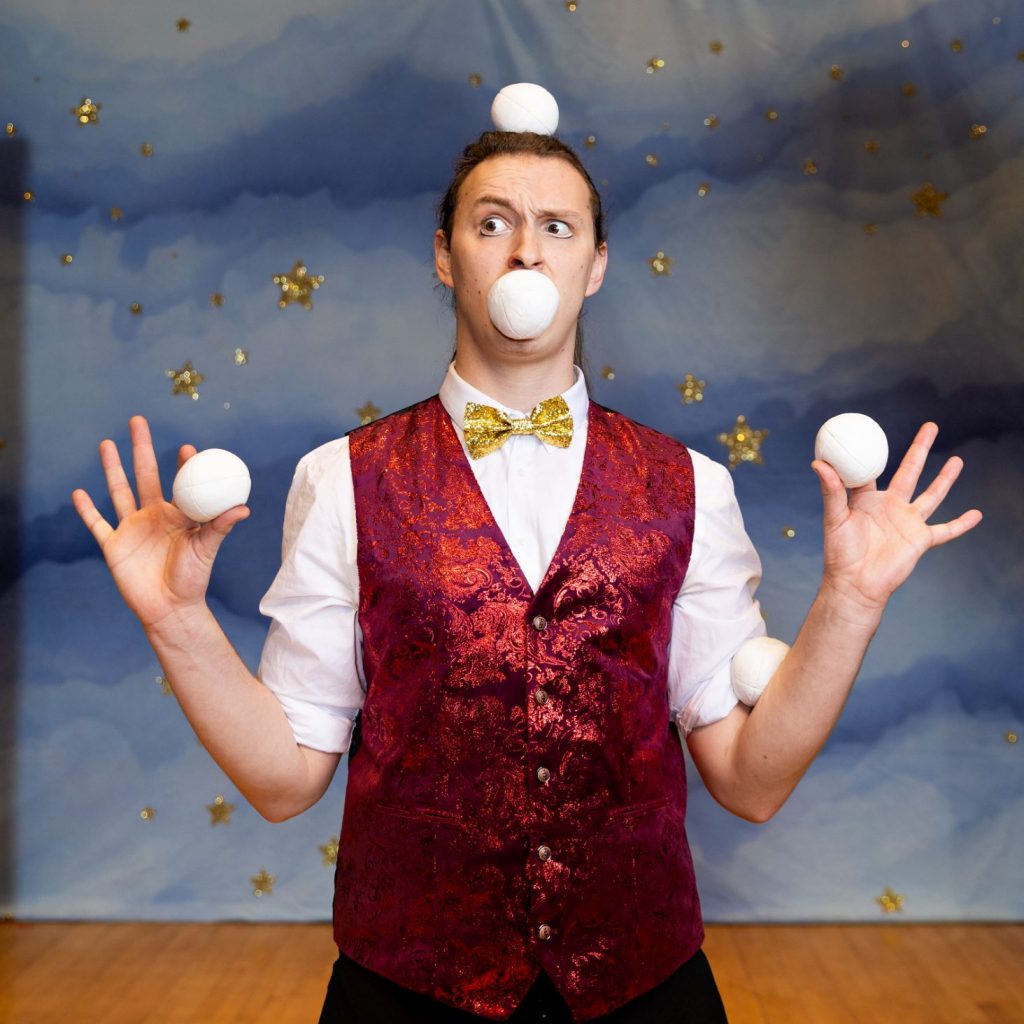 A man is wearing a gold bow tie, he is holding juggling balls in his hand and mouth, he even has one on top of this head