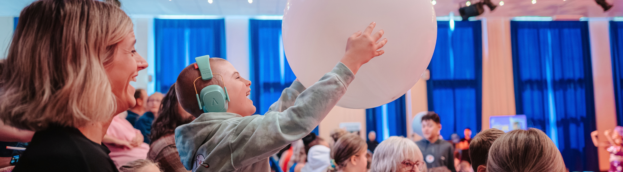 A young boy is wearing ear defenders, he is smiling as he reaches out for a balloon that has been thrown in the audience.