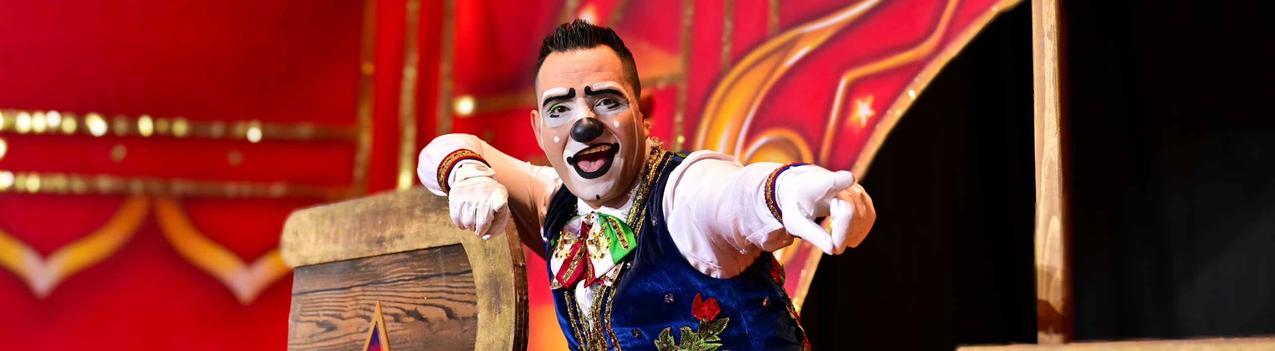 A clown has an excited smile and points towards the camera. He is wearing white gloves, a blue waistcoat and a colourful bow tie.