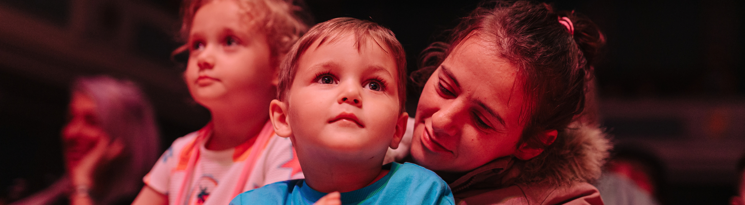 A white boy is mesmerised as he looks towards the stage. He has brown hair and is sitting on his mums lap.