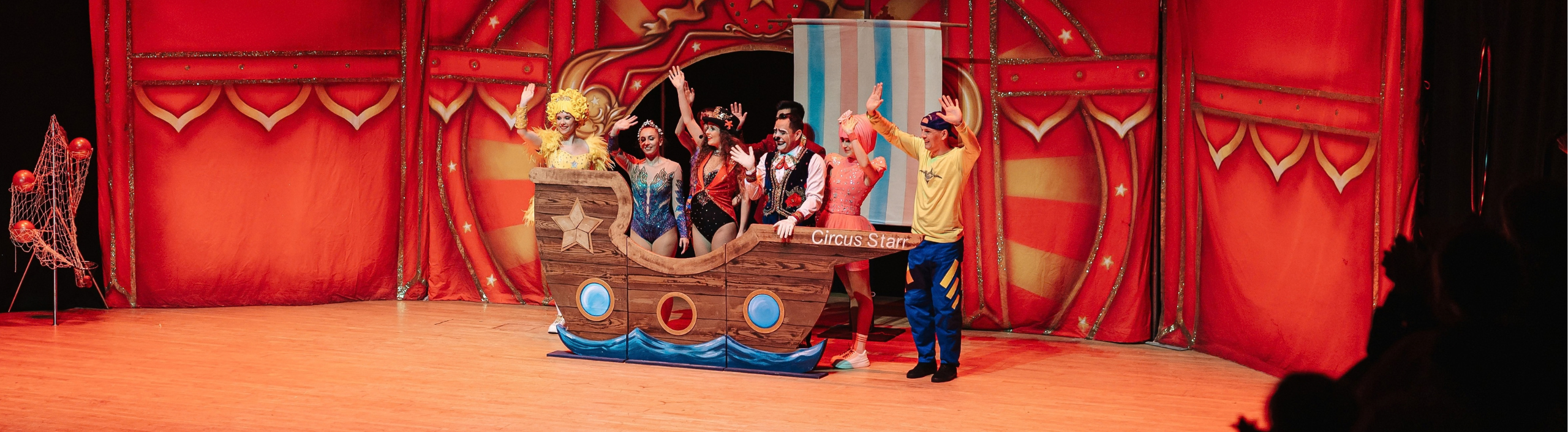The Circus Starr troupe are waving at the audience while they stand on stage. They are standing behind a wooden ship.