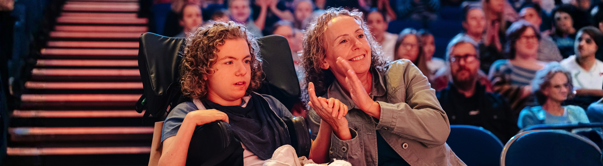 A young person is sat in a wheelchair looking at the stage. They have curly brown hair and are holding hands with the woman who is sat next to them. She is smiling and clapping as she watches the show.
