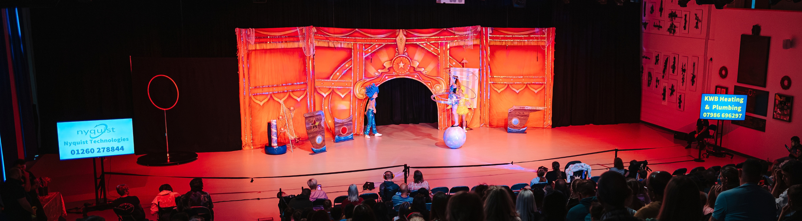 The Circus Starr backdrop is red with gold glitter. There is a girl performing with hula hoops as she balances on a ball. There are two screens either side of the stage promoting business information.