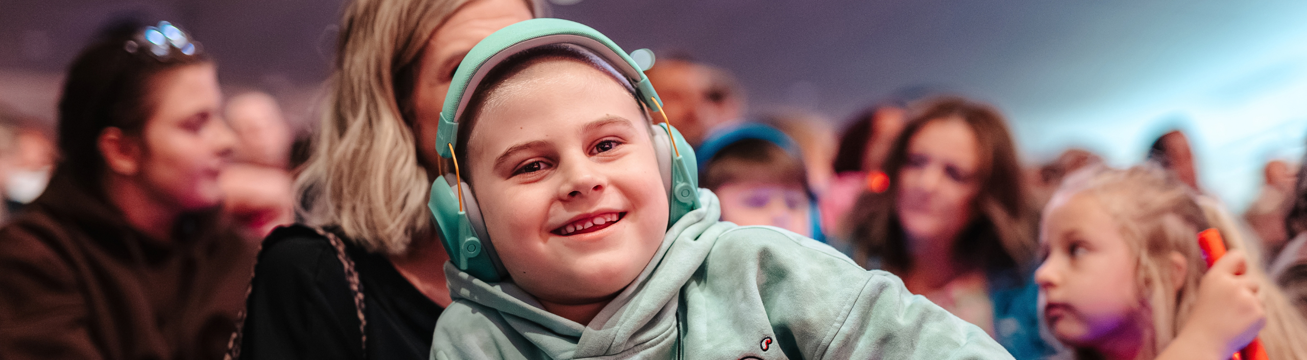 A white boy is smiling in the audience, He is wearing a mint coloured hoody and matching ear defenders.