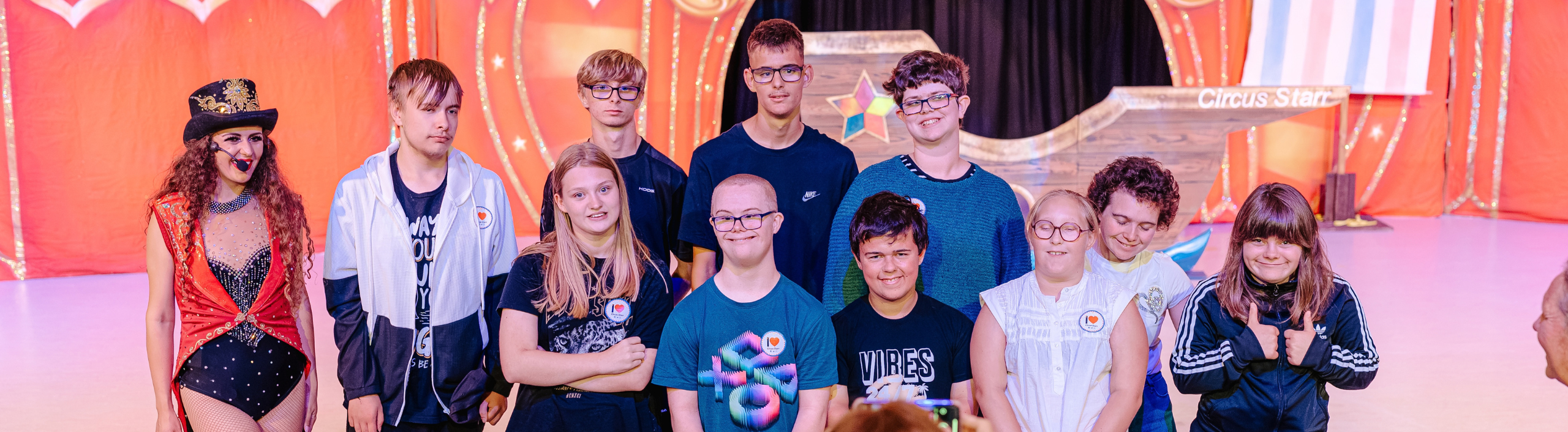 A large group of children are standing in front of the stage and posing for a picture with the ringmistress.
