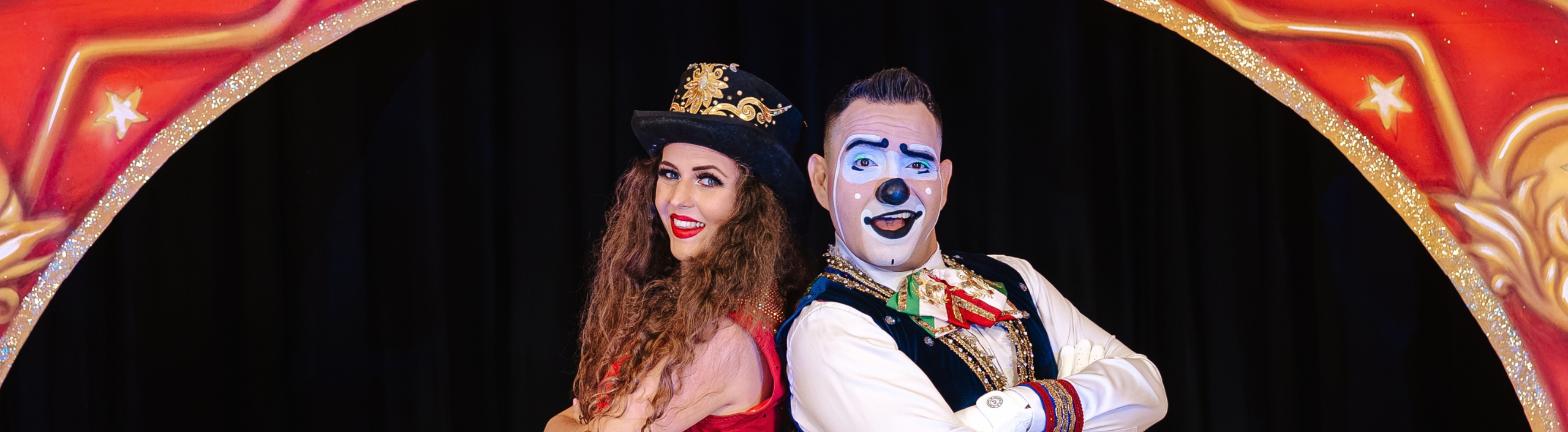 A ring mistress and a clown are stood back to back and are turning to towards the camera. They both have their arms crossed and are smiling towards the camera, the clown has an especially cheeky grin!