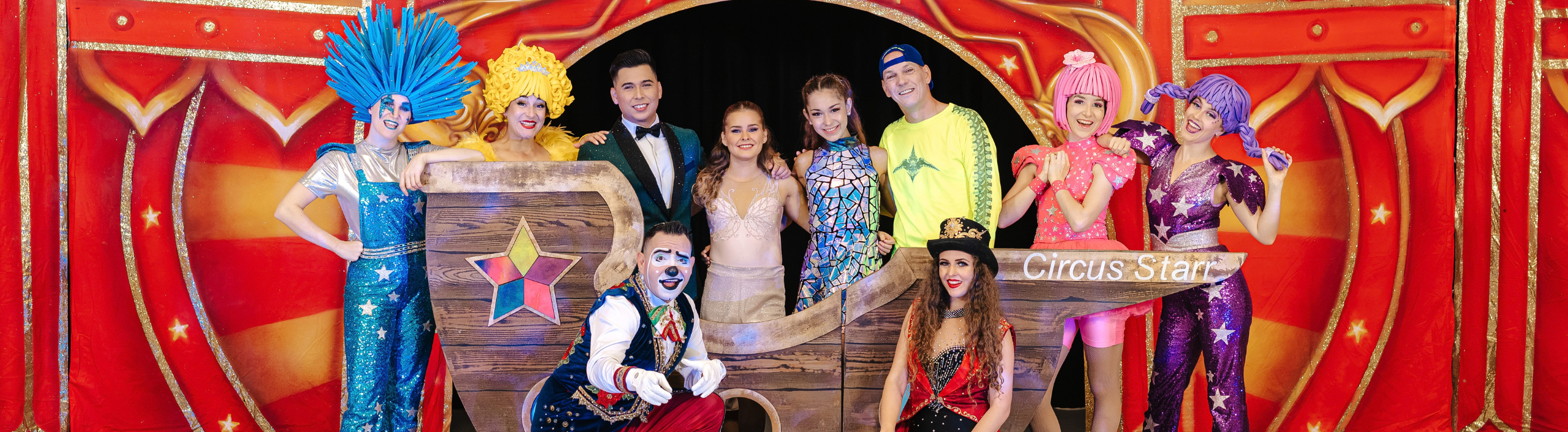 The circus troupe pose on stage, they are smiling at the camera. They are wearing colourful sparkly clothing and make up.