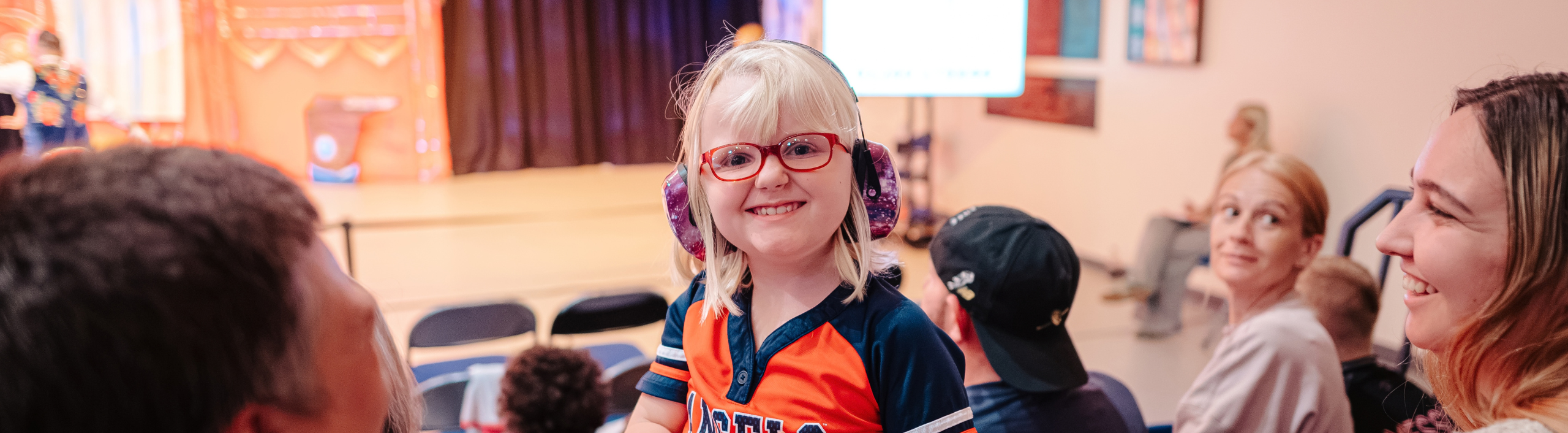 A little white girl is smiling at the camera as she stands in the audience. She has red glasses, blonde hair, and purple ear defenders.