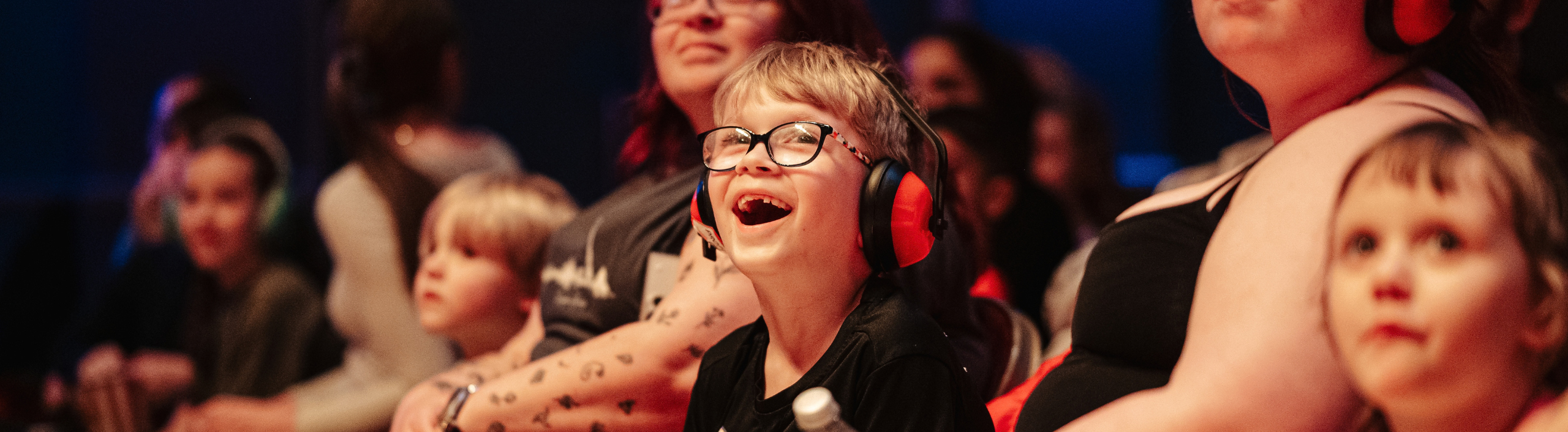 A little boy sits in the audience, he has a very excited smile on his face. He is wearing glasses and ear defenders.