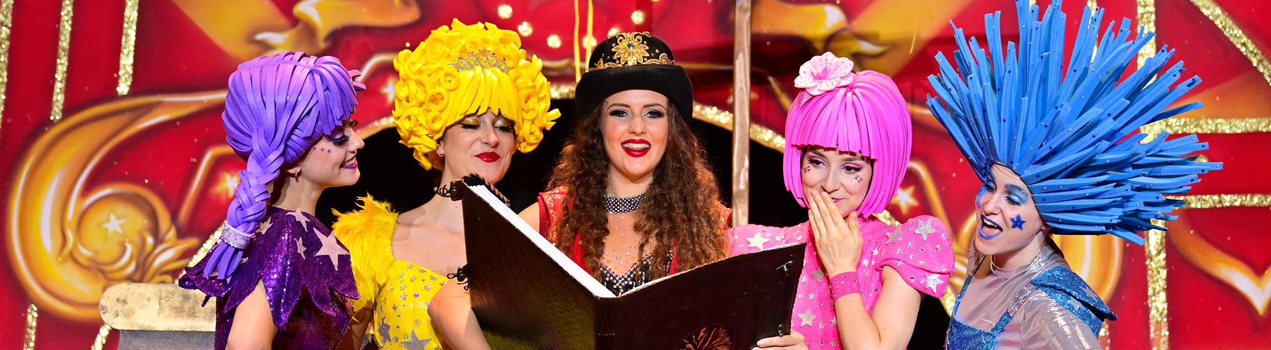 The Ringmistress stands in the middle of a The Starr Squad, who are dressed in colourful outfits. The ringmistress has a sparkly top hat and is smiling as she holds a large book.